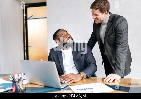 Happy business people discussing work questions in office, using laptop, developing a new project together. African american male employee asking an advice from caucasian man colleague, smiling Stock Photo
