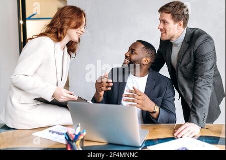 Happy friendly coworkers sitting in office, having a pleasent conversation. Multiracial business people talking about work process, smiling, discussing ideas for new startup Stock Photo
