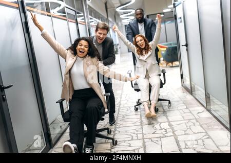 Joyful male and female colleagues take a break from work, having fun. Happy multiracial office employees in formal wear making a race on chairs, smiling and laughing Stock Photo