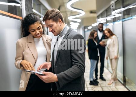Multiethnic coworkers are standing in office, looking at the tablet. Caucasian male employee asking an advice from beautiful african american female colleague about some work problems Stock Photo