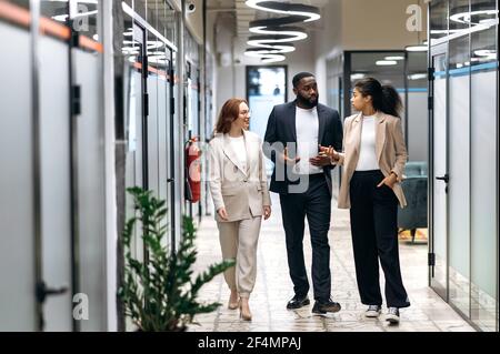 Concentrated multiracial colleagues in formal wear walking in office hall, discussing the project. Young adult business partners consulting each other on work questions, communication concept Stock Photo