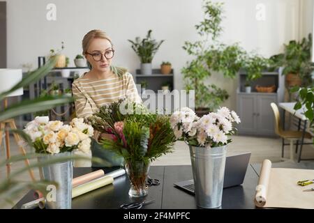 Portrait of young blonde woman arranging bouquets while working in green florists workshop, copy space Stock Photo
