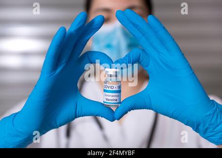 Close up of unrecognizable nurse with medical mask and protective gloves. She is forming a heart with her hands showing a glass vial of Covid-19 vacci Stock Photo