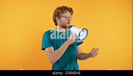redhead man in glasses holding megaphone isolated on yellow Stock Photo