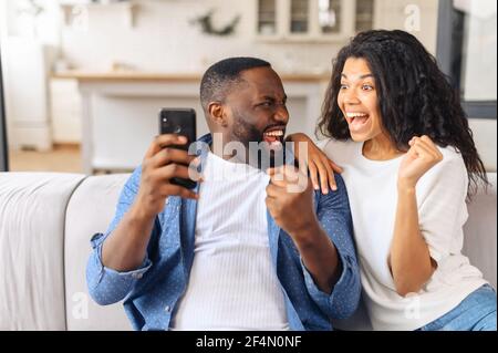 Excited and overjoyed African-American young couple sits on the couch at home and holds smartphone, a man and woman show victory gesture, celebrating good news or win, received money on account Stock Photo