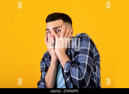 Millennial man feeling scared, trying to hide, covering his face with hands on orange studio background Stock Photo