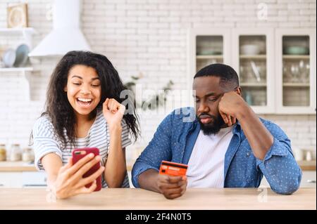 Excited from long awaited purchase multi ethnic woman holds smartphone and rejoices while sad greedy African-American man sitting near with a credit card, not happy to spend money Stock Photo
