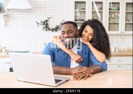 Cute multiracial couple enjoys leisure time at home with a laptop, happy woman and man in embrace look at the screen and laughs, watching videos, video calling with friends Stock Photo