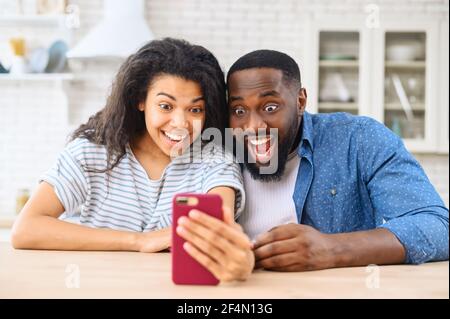 Amazed African-American couple looks at smartphone screen, surprised man and woman staring with wide opened eyes, received job offer, impressed with news feed in social networks Stock Photo