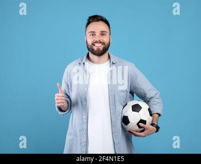 Cheerful young guy holding soccer ball, making thumb up gesture, recommending sports on blue studio background Stock Photo