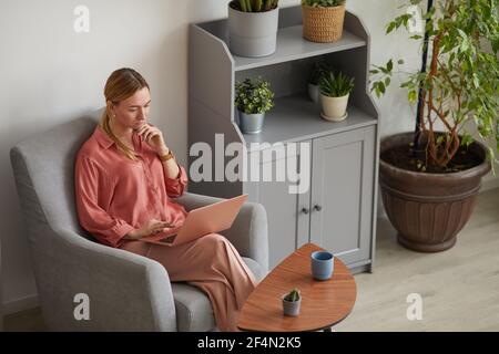Minimal high angle portrait of modern successful businesswoman using laptop and working while sitting in armchair in stylish home interior, copy space Stock Photo