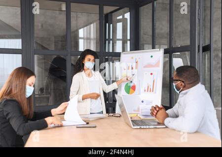 Diverse work team in protective medical masks working together in modern coworking space, multiracial employee brainstorming during pandemic period, a biracial woman presenting ideas. New social rules Stock Photo