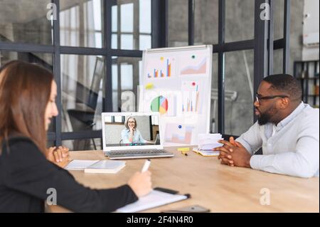 Colleagues have video meeting in the office, young coworkers sit at the desk and communicating with senior female via video call on the laptop, conference with a leader online, briefing Stock Photo