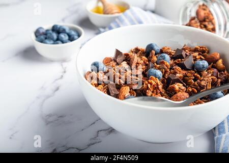 Homemade granola, muesli with pieces of dark chocolate, nuts and blueberries in bowl on white marble background. Healthy breakfast. Selective focus. Stock Photo