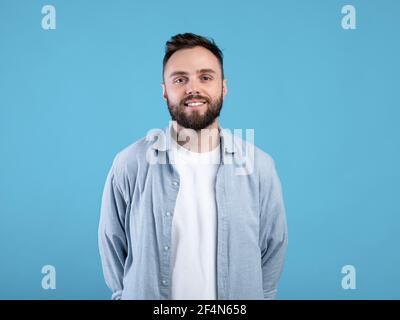 Portrait of cheerful young guy in casual wear posing and smiling at camera on blue studio background Stock Photo