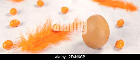 Banner with Egg on a white concrete background with orange feathers and orange chocolates in the shape of an egg. Happy Easter concept. Stock Photo
