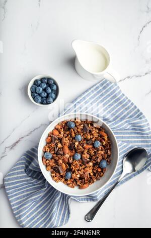 Homemade chocolate granola, muesli with almonds, hazelnuts and blueberries in bowl on white marble background. Healthy breakfast. Top view. Flat lay m Stock Photo