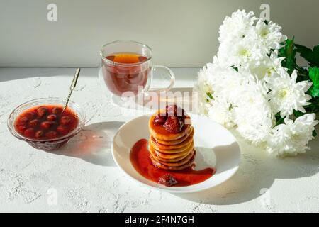 Fritters in a white plate with strawberry jam, a cup of tea and a bouquet of white chrysanthemums on a white table. Stock Photo