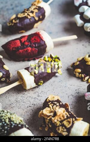 Bananas in dark chocolate with different toppings on stick, gray background. Healthy vegan dessert concept. Stock Photo