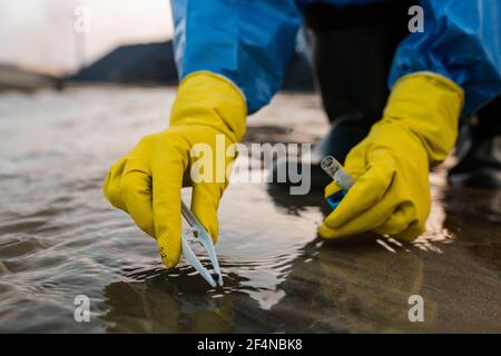 Contemporary female ecologist in rubber gloves and protective blue coveralls sitting on squats while taking sample of toxic water Stock Photo