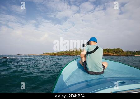 Photographer sitting on boat. Young man photographing beautiful coast with palm tree near Tangalle in Sri Lanka. Stock Photo