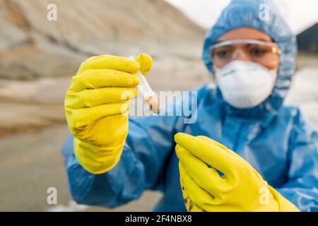 Gloved hands of young female ecologist in respirator and eyeglasses holding flask with sample of dirty water from polluted and toxic area Stock Photo