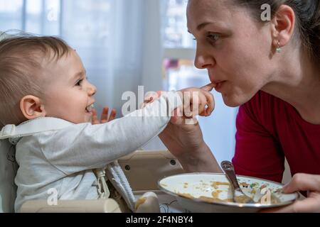 Happy and messy little baby and his mother feed each other. Healthy eating education since early days. Stock Photo