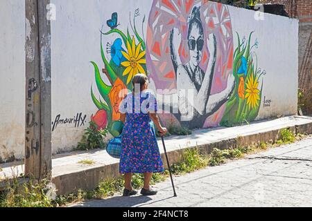 Elderly Mexican woman walking with cane past graffiti street art on wall in the city San Miguel de Allende, Guanajuato, Central Mexico Stock Photo