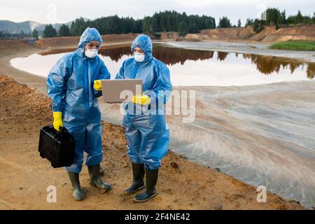 Two young female ecologists in protective workwear standing on polluted soil or clay against lake with dirty water and making notes in laptop Stock Photo