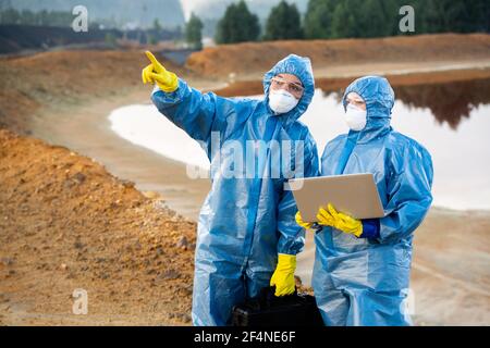 Young female researcher pointing forwards while showing colleague with laptop where they should take samples of toxic soil and water Stock Photo