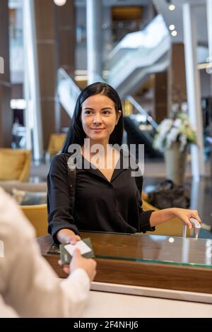 Happy young businesswoman or female traveler with luggage standing by reception counter and passing her documents to receptionist Stock Photo