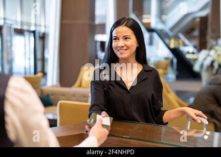 Happy young businesswoman or female traveler with luggage passing her documents to hotel receptionist over counter before registration Stock Photo