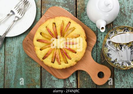 Homemade round cake with apple slices looking like sun rays with teaware on old wooden blue table above view Stock Photo
