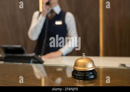 Bell on wooden reception counter against female receptionist consulting clients on the phone while working in contemporary luxurious hotel Stock Photo