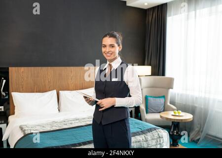 Happy young female worker of luxurious hotel in uniform standing in front of camera against bed and using touchpad while looking at you Stock Photo