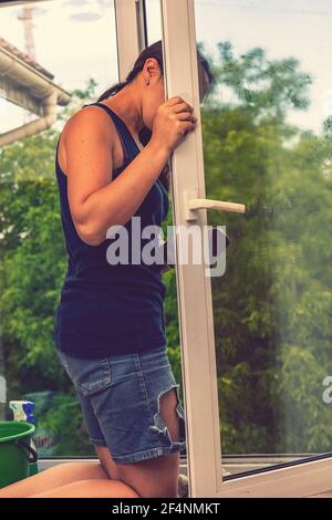 Beautiful woman cleaning window at home. A girl washes windows at home. To clean up the house. Long-haired woman cleaning windows with spray in home Stock Photo