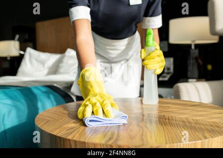 Young contemporary chambermaid in uniform and protective gloves using duster and detergent while cleaning wooden table in hotel room Stock Photo