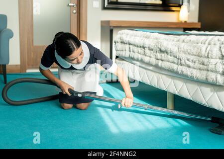 Young beautiful brunette chambermaid in uniform using vacuum cleaner while cleaning blue floor covering under bed in hotel room Stock Photo