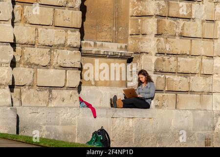 Oxford, UK 22 Nov 2020: Student girl sitting in the sun on Radcliffe Camera wall slope using laptop for learning. Young woman research information on Stock Photo