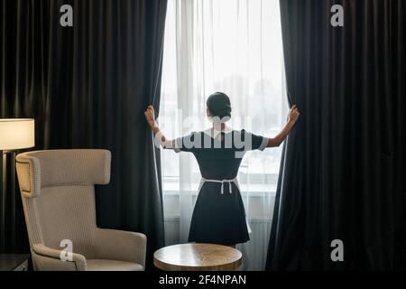 Back view of young chambermaid in uniform opening dark curtains hanging on large window of hotel room before cleaning it Stock Photo