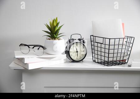 A stack of books, an artificial succulent plant in a ceramic pot, glasses, a metal basket with notebooks, a pen are on a white table surface. Concept Stock Photo
