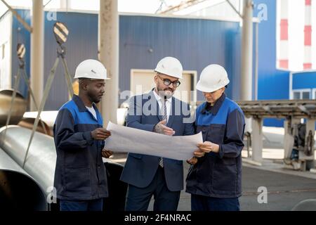 Group of three young contemporary workers of industrial plant discussing unfolded blueprint with sketches of new equipment in workshop Stock Photo