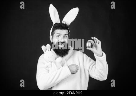 Paschal egg. Comic hipster with rabbit ears holding bunny toy and egg. Bearded man in rabbit costume with easter egg and hare toy. This easter egg is Stock Photo