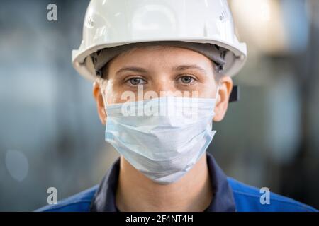 Young female factory worker in overalls, hardhat and protective mask looking at you while standing in workshop in front of camera Stock Photo