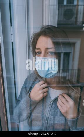 Young depressed unhappy caucasian woman with face mask looking worried and lonely through the window in quarantine at home. Feeling worthless and in p Stock Photo