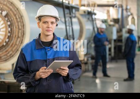 Young female factory worker in overalls and hardhat looking at you while standing in front of camera in workshop and using digital tablet Stock Photo