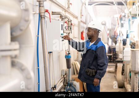 Young African male worker in overalls and hardhat standing in front of huge industrial machine and turning knob on control panel Stock Photo