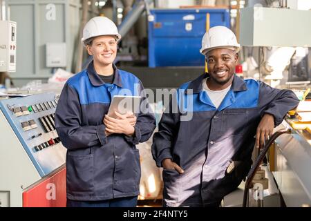 Two young successful workers of modern plant in overalls and hardhats standing in front of camera against huge industrial machine in workshop Stock Photo