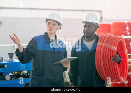 Young contemporary female worker of modern plant using tablet and showing her African male colleague new industrial machine Stock Photo