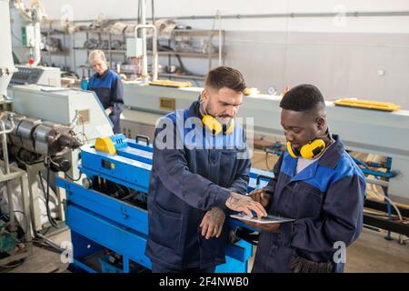 Two young intercultural male workers of modern plant discussing online technical data while standing by industrial machine in workshop Stock Photo